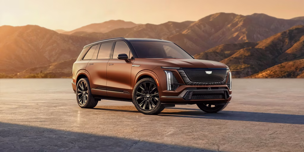 2027-cadillac-vistiq-mov-limited-editions-adobe-frost-2000x1000-27cdvist070002 | Wagner Certified Collision Bronze luxury SUV parked on a flat desert surface with orange sunset and distant mountains behind it, panoramic scene.