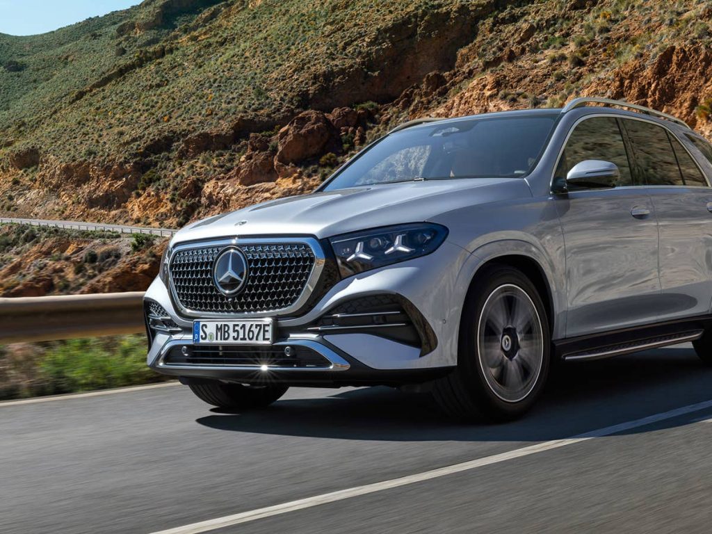 Silver Mercedes-Benz SUV driving along a winding coastal mountain road, rocky hills in the background. It’s in motion, front three-quarter view.] ,