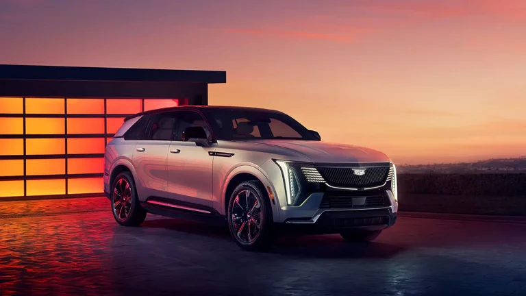 Silver Cadillac SUV parked outdoors at sunset, with a modern glass-wall building reflecting pink-orange sky behind it.