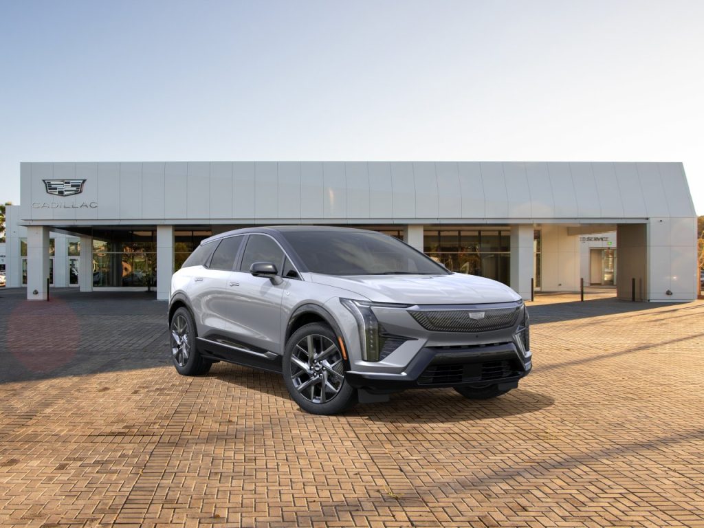 fa5a5db | Wagner Certified Collision Silver Cadillac SUV parked on a brick lot in front of a modern Cadillac dealership building with logo visible on the facade, daytime light