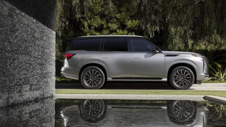 Silver luxury SUV parked on a driveway beside a stone wall and lush greenery, its reflection visible in the water feature nearby.